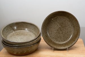 the photograph shows 4 clay coyote flameware mini savory pie dishes on a small light colored wooden table. there is a stack of three flameware mini savory pie dishes on the left side of the small table. they are stacked on ontop of the other nesting style. on the right side of the small table there is one flameware mini savory pie dish that is in a stand, allowing it to be almost vertical. the mini savory in the stand has its entire cooking surface visible due to this. the mini savory pie dishes are glazed in coyote grey (grey with dark speckling through out). the background is a white wall. the photograph is lit with white light.