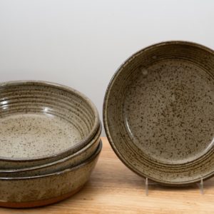 the photograph shows 4 clay coyote flameware mini savory pie dishes on a small light colored wooden table. there is a stack of three flameware mini savory pie dishes on the left side of the small table. they are stacked on ontop of the other nesting style. on the right side of the small table there is one flameware mini savory pie dish that is in a stand, allowing it to be almost vertical. the mini savory in the stand has its entire cooking surface visible due to this. the mini savory pie dishes are glazed in coyote grey (grey with dark speckling through out). the background is a white wall. the photograph is lit with white light.