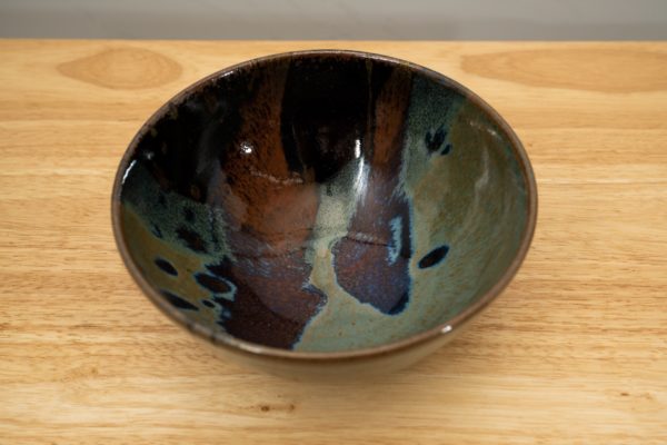 the photograph shows a high angle view of a clay coyote soup and chili bowl glazed in zappa. the soup and chili bowl is resting on a light colored wooden surface. the angle of the photograph is high enough to allow the viewer a look at the inside of the bowl, and the zappa design contained within it. the photograph is lit with white light.