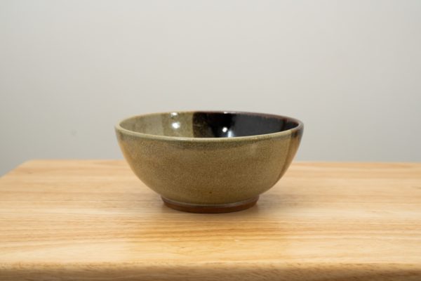 the photograph shows a clay coyote soup and chili bowl glazed in mint chip resting on a small light colored wooden table. the background is a white wall. the photograph is well lit with white light.