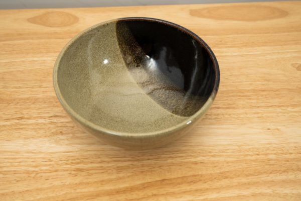the photograph shows a high angle view of a clay coyote soup and chili bowl. the photograph is taken from a high angle, to allow the viewer to see the mint chip design on the inside of the bowl. the bowl is resting on a light colored wooden table. the photograph is lit with white light.