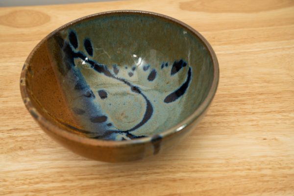 the photograph shows a high angle view of a clay coyote soup and chili bowl resting on a light colored wooden surface. the photographs angle is at a height that allows the viewer to see the joes blue pattern on the inside of the bowl. the photograph is lit with white light.
