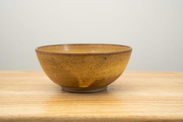 the photograph shows a clay coyote chili and soup bowl glazed in yellow salt. the bowl is resting flat on a light colored wooden surface. the photograph is lit with white light.