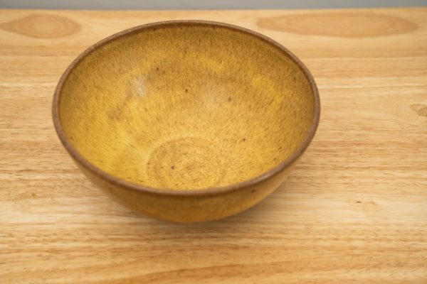 the photograph shows a high angle view of a clay coyote soup and chili bowl glazed in yellow salt. the bowl is resting on a light colored wooden table. the photograph is lit with white light.