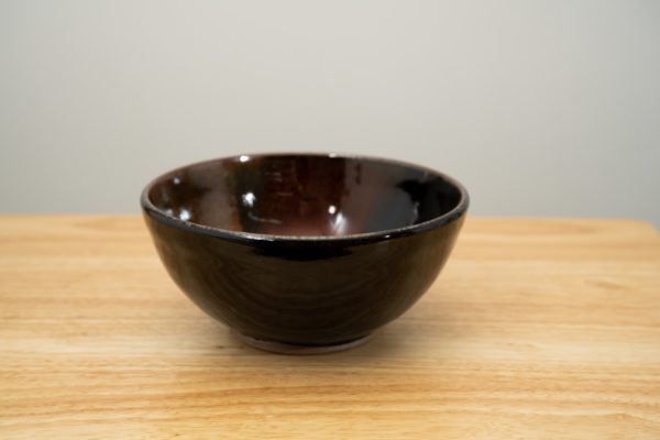 the photograph shows a clay coyote soup and chili bowl glazed in merlot resting flat on a light colored wooden table. the background is a plain white wall. the photograph is lit with white light.