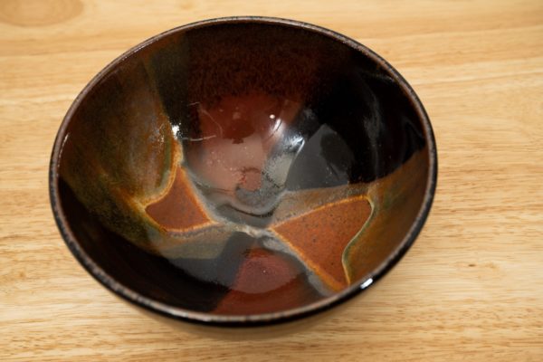 the photograph shows a high angle view of a clay coyote soup and chili bowl glazed in merlot. the angle of the photograph allows the viewer to see the merlot pattern on the inside of the bowl. the bowl is resting on a light colored wooden surface. the photograph is lit with white light.