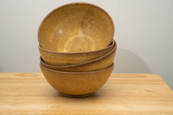 the photograph shows a stack of 4 clay coyote soup and chili bowls glazed in yellow salt all stacked on top of each other. the bowls are resting on a small light colored wooden table. the background is a white painted wall. the photograph is lit with white light.