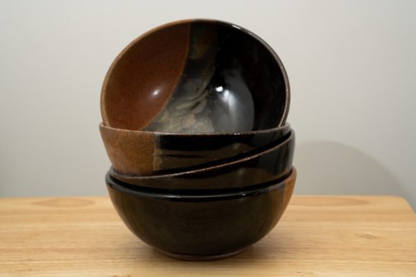 the photograph shows a stack of 4 clay coyote soup and chili bowls all glazed in mocha swirl. they are stacked one on top of the other, with the top bowl resting slightly on its side to provide a look at the glaze pattern on the inside. the stack of bowls is resting on a small light colored wooden table. the background is a plain white wall.