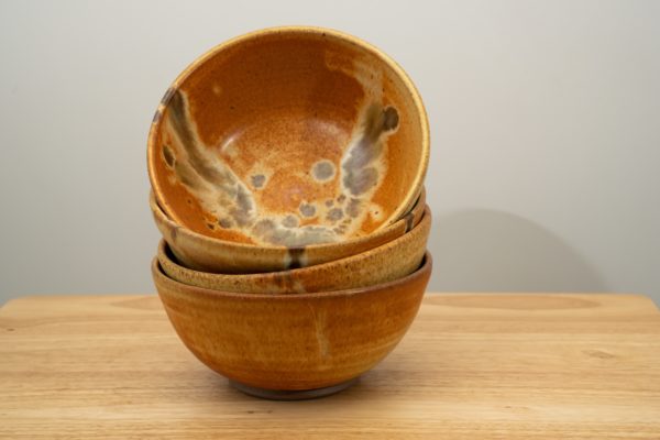 the photograph shows a stack of 4 clay coyote bowls resting on a light colored wooden table. the top bowl of the stack has been tipped to its side to allow the viewer a good look at the feather pattern on the inside of the bowl. the bowls are resting on a small light colored wooden table. the background is a white wall. the photograph is well lit with white light.