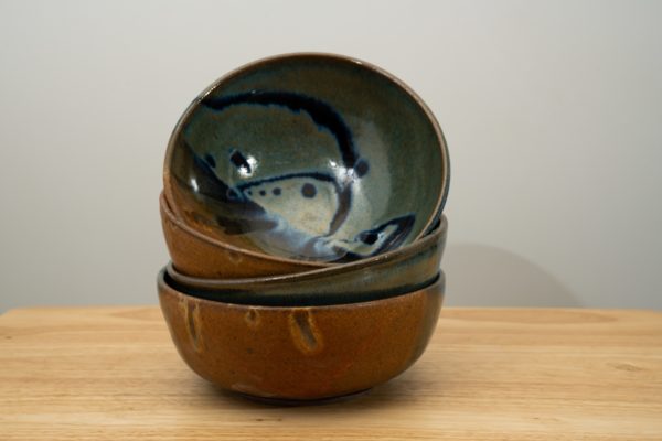 the photograph shows a stack of 4 clay coyote soup and chili bowls stack on top of each other. the top bowl of the stack of 4 is resting on its side to allow a good view of the joes blue pattern on the inside of the bowl. the stack of bowls is resting on a small light colored wooden table. the background is a white wall. the photograph is lit with white light.