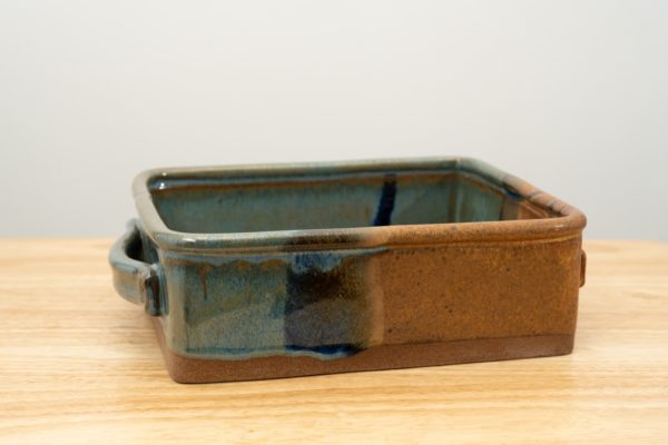 the photograph shows a clay coyote baking dish glazed in joes blue resting on a light colored wooden surface. the front right corner of the baking dish is the brown section of the joes blue glaze pattern. the background is a plain white wall. the photograph is well lit with white light.