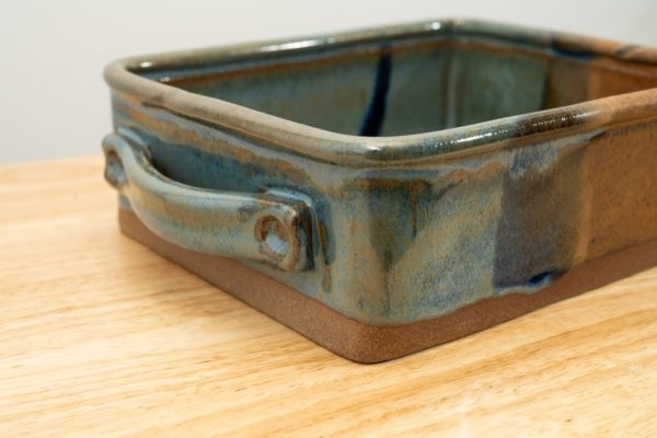 the photograph shows a clay coyote baking dish glazed in joes blue resting on a light colored wooden surface. the front right corner of the baking dish is the brown section of the joes blue glaze pattern. the background is a plain white wall. the photograph is well lit with white light.