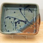 the photograph shows a clay coyote baking dish glazed in joes blue resting on its side with the help of a clear plastic holder. the baking dish is resting on a small light colored wooden table. the baking dish is resting on its side, allowing for full view of the glaze pattern inside the baking dish. the lower right corner is the brown corner of the joes blue pattern. the background is a white wall. the photograph is well lit with white light.