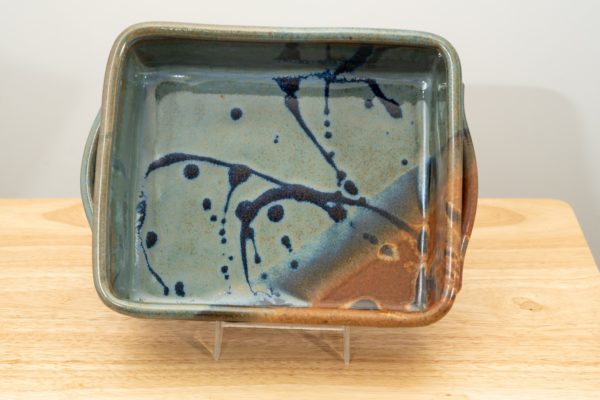 the photograph shows a clay coyote baking dish glazed in joes blue resting on its side with the help of a clear plastic holder. the baking dish is resting on a small light colored wooden table. the baking dish is resting on its side, allowing for full view of the glaze pattern inside the baking dish. the lower right corner is the brown corner of the joes blue pattern. the background is a white wall. the photograph is well lit with white light.