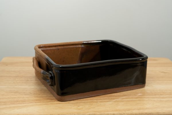 the photograph shows a clay coyote baking dish glazed in mocha swirl resting on a small light colored wooden table. the brown part of the mocha swirl pattern is in the back left corner of the dish. the background is a white wall. the photograph is lit with white light.