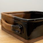 the photograph shows a clay coyote baking dish glazed in mocha swirl resting on a small light colored wooden table. the brown part of the mocha swirl pattern is in the back left corner of the dish. the background is a white wall. the photograph is lit with white light.