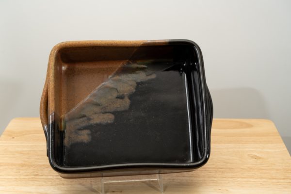 the photograph shows a clay coyote baking dish glazed in mocha swirl resting on its side to show off the glaze pattern on the inside of the baking dish. the upper left corner is the brown section of the mocha swirl pattern. the baking dish is resting on its side with the help of a small clear plastic stand. the baking dish is resting on a small light colored wooden table. the table is sitting in front of a plain white wall. the photograph is lit with white light.
