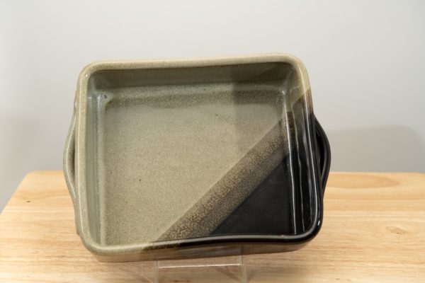 the photograph shows a clay coyote baking dish glazed in mint chip resting on its side. the baking dish is resting on its side with the help of a small clear plastic stand. the position of the baking dish allows for the viewer to see the inside of the baking dish and the glaze pattern it contains. the lower right corner is the black part of the mint chip pattern. the other three corners are mint colored. the background is a white wall. the photograph is lit with white light.