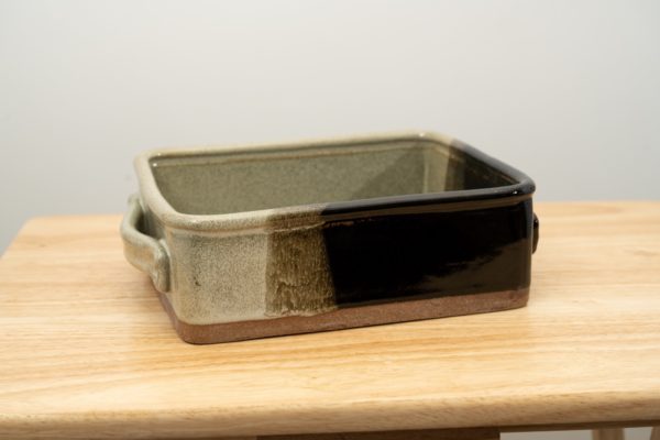 the photograph shows a clay coyote baking dish glazed in mint chip. the dark corner of the mint chip pattern is in the front right corner of the baking dish. the baking dish is resting on a small light colored wooden table. the table and dish is front of a white wall. the photograph is well lit with white light.