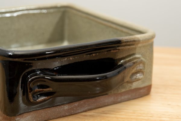 the photograph shows the handle end of a clay coyote baking dish glazed in mint chip. the dark part of the glaze pattern is the closest corner to the viewer, and covers the handle that is the main focus of the photograph. the baking dish is resting on a light colored wooden surface. the background is a white wall.