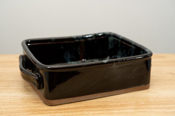 the photograph shows a clay coyote baking dish glazed in midnight garden resting on a small light colored wooden table. the main part of the midnight garden color pattern is in the back right corner, furthest from the camera. the background is a white wall. the photograph is lit with white light.