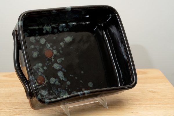 the photograph shows a clay coyote baking dish glazed in midnight garden resting on its side. the baking dish is resting on its side with the help of a small clear plastic stand. the position of the baking dish allows for the viewer to see the inside of the baking dish and the glaze pattern it contains. the background is a white wall. the photograph is lit with white light.