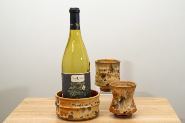 the photograph shows the clay coyote date night set. the date night set consists of two yunomis and a wine bottle coaster. in the photograph starting from the left, the wine bottle coaster has a demonstration empty wine bottle in it and is glazed in feather. to the right and slightly closer to the camera one of the yunomis, also glazed in feather. almost directly behind the first yunomi is the second, also glazed in feather. it is resting on a small clear stand allowing it to be almost completely visible despite being "behind" the first. the date night set is resting on a small light colored wooden table. the background is a white wall. the photograph is well lit with white light.