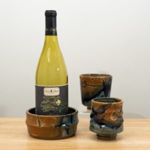 the photograph shows the clay coyote date night set. the date night set consists of two yunomis and a wine bottle coaster. in the photograph starting from the left, the wine bottle coaster has a demonstration empty wine bottle in it and is glazed in joes blue. to the right and slightly closer to the camera one of the yunomis, also glazed in joes blue. almost directly behind the first yunomi is the second, also glazed in joes blue. it is resting on a small clear stand allowing it to be almost completely visible despite being "behind" the first. the date night set is resting on a small light colored wooden table. the background is a white wall. the photograph is well lit with white light.