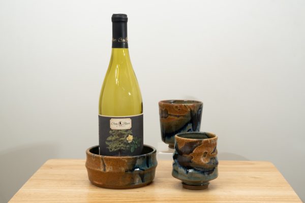 the photograph shows the clay coyote date night set. the date night set consists of two yunomis and a wine bottle coaster. in the photograph starting from the left, the wine bottle coaster has a demonstration empty wine bottle in it and is glazed in joes blue. to the right and slightly closer to the camera one of the yunomis, also glazed in joes blue. almost directly behind the first yunomi is the second, also glazed in joes blue. it is resting on a small clear stand allowing it to be almost completely visible despite being "behind" the first. the date night set is resting on a small light colored wooden table. the background is a white wall. the photograph is well lit with white light.
