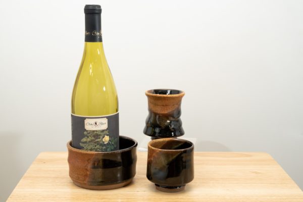 the photograph shows the clay coyote date night set. the date night set consists of two yunomis and a wine bottle coaster. in the photograph starting from the left, the wine bottle coaster has a demonstration empty wine bottle in it and is glazed in mocha swirl. to the right and slightly closer to the camera one of the yunomis, also glazed in mocha swirl. almost directly behind the first yunomi is the second, also glazed in mocha swirl. it is resting on a small clear stand allowing it to be almost completely visible despite being "behind" the first. the date night set is resting on a small light colored wooden table. the background is a white wall. the photograph is well lit with white light.