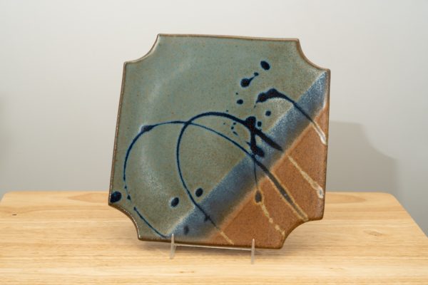 the photograph shows a clay coyote sushi plate glazed in joes blue standing upright with the help of a small clear plastic stand. the plate is resting on a small light colored wooden table. the background is a white wall. the photograph is lit with white light.