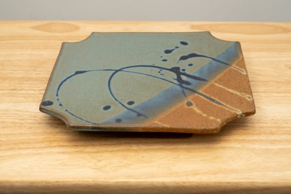 the photograph shows a clay coyote sushi plate glazed in joes blue. the sushi plate is resting on a light colored wooden surface. the brown part of the glaze pattern is in the lower right corner of the plate.