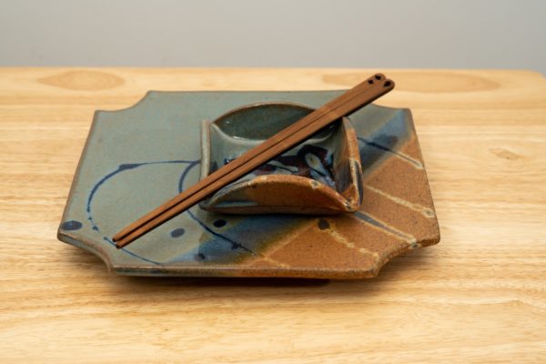 the photograph shows a clay coyote sushi plate and little dipper glazed in joes blue. the little dipper is the square style. the little dipper is resting on top of the sushi plate. on top of the little dipper is a pair of chopsticks. the chopsticks are moonspoon brand, which are sold at the clay coyote gallery. the tips of the chopsticks are pointed towards the lower left and the tops are pointed towards the upper right. the whole stack is resting on a small light colored wooden table. the background is a white wall. the photograph is well lit with white light.