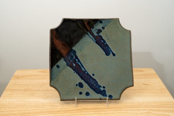 the photograph shows a clay coyote sushi plate standing upright with the help of a small clear plastic stand. the dark part of the zappa glaze is located on the upper left of this plate. the plate is resting on a small light colored wooden table. the background is a white wall. the photograph is lit with white light.
