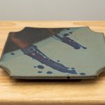 the photograph shows a clay coyote sushi plate glazed in zappa. the sushi plate is resting flat on a light colored wooden table. the background is a white wall. the darkest part of the glaze is in the upper left corner of the plate.