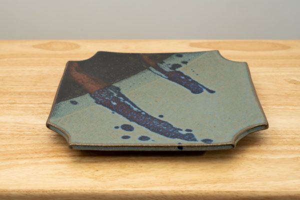the photograph shows a clay coyote sushi plate glazed in zappa. the sushi plate is resting flat on a light colored wooden table. the background is a white wall. the darkest part of the glaze is in the upper left corner of the plate.