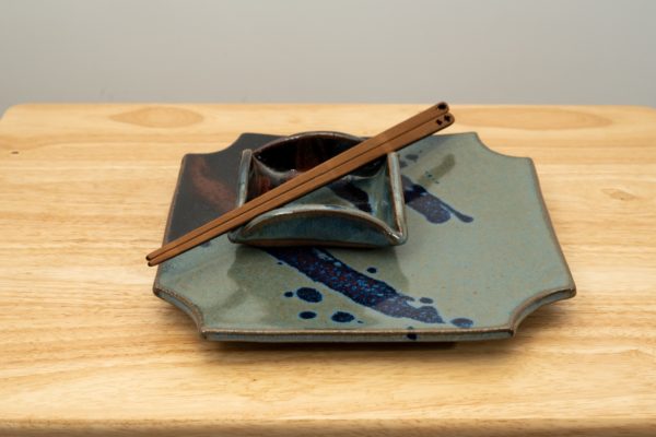 the photograph shows a clay coyote sushi plate and little dipper glazed in zappa. the little dipper is the square style. the little dipper is resting on top of the sushi plate. on top of the little dipper is a pair of chopsticks. the chopsticks are moonspoon brand, which are sold at the clay coyote gallery. the tips of the chopsticks are pointed towards the lower left and the tops are pointed towards the upper right. the whole stack is resting on a small light colored wooden table. the background is a white wall. the photograph is well lit with white light.
