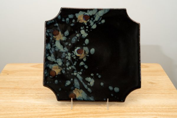 the photograph shows a clay coyote sushi plate glazed in midnight garden standing upright with the help of a small clear plastic stand. the colorful part of the midnight design is on the left side of the plate. the plate is resting on a small light colored wooden table. the background is a white wall. the photograph is lit with white light.