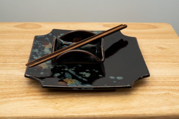 the photograph shows a clay coyote sushi plate and little dipper both glazed in midnight garden. the little dipper is resting on top of the sushi plate. the little dipper is the square style. the little dipper has a pair of moonspoon chopsticks resting on top of them. the tips of the chopsticks are pointing towards the lower left corner and the tops towards the upper right. the stack of the sushi plate with little dipper and chopsticks on top are all resting on a small light colored wooden table. the background is a white wall.