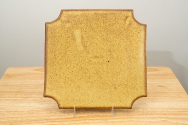 the photograph shows a clay coyote sushi plate glazed in yellow salt standing with the help of a small clear plastic stand. the plate and stand are resting on a small light colored wooden table. the background is a white wall. the photograph is lit with white light.