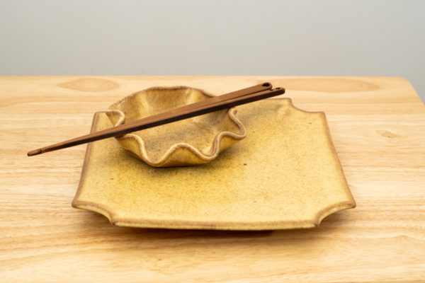 the photograph shows a clay coyote sushi plate glazed in yellow salt with a clay coyote little dipper also glazed in yellow salt resting on top of it. on top of the little dipper there is a pair of moonspoon chopsticks (sold in the clay coyote gallery). the chopsticks are resting diagonally on the little dipper, with the top of the chopsticks pointed towards the upper right corner, and the tips pointed towards the lower left of the photograph. the stack of the sushi plate, little dipper and chopsticks are resting on a light colored wooden table. the background is a white wall. the photograph is well lit with white light.