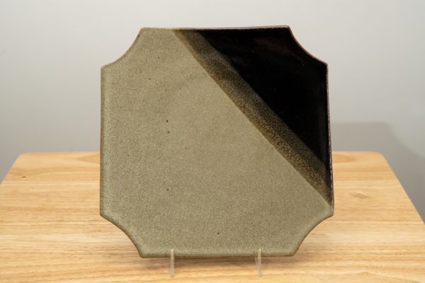 the photograph shows a clay coyote sushi plate glazed in mint chip standing almost vertical with the help of a small clear plastic stand. the dark part of the glaze pattern is in the top right corner of the plate. the plate is resting on a small light colored wooden table. the background is a white wall. the photograph is lit with white light.