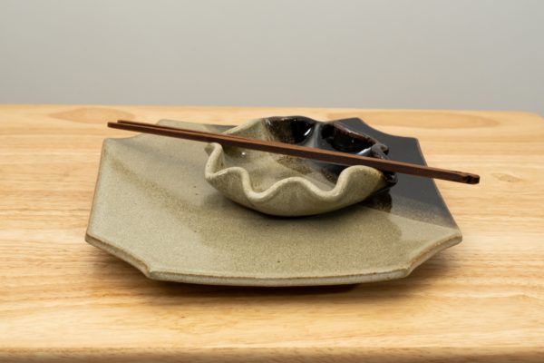 the photograph shows a clay coyote sushi plate with a clay coyote little dipper resting on top of it. both pieces are glazed in mint chip. both pieces have the black part of the glaze pattern in the back right corner in the photograph. on top of the little dipper is a pair of moonspoon chopsticks (sold in the clay coyote gallery). the stack of the sushi plate, little dipper and moonspoon chopsticks are all resting on a light colored wooden table. the background is a white wall. the photograph is lit with white light.