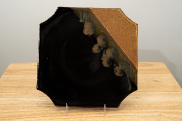 the photograph shows a clay coyote sushi plate glazed in mocha swirl. the plate is sitting near vertically with the help of a clear plastic stand. the brown part of the mocha swirl pattern is in the upper right corner of the photograph. the sushi plate and stand are resting on a small light colored wooden table. the background is a white wall.