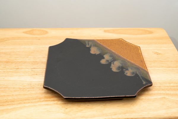 the photograph shows a clay coyote sushi plate glazed in mocha swirl sitting flat on a small light colored wooden table. the brown part of the mocha glaze pattern is in the back right corner of the plate. the background is a white wall. the photograph is lit with white light.