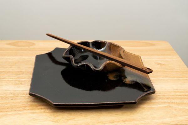 the photograph shows a 2 clay coyote pottery pieces and a pair of moonspoon (sold at clay coyote) chopsticks. the sushi plate and little dipper are both glazed in mocha swirl. the little dipper is sitting on top of the sushi plate. the moonspoon chopsticks are laying across the top of the little dipper diagonally. the top of the chopsticks are pointed towards the lower right and the tips are pointed towards the upper left. the background is a white wall. the photograph is lit with neutral white light.