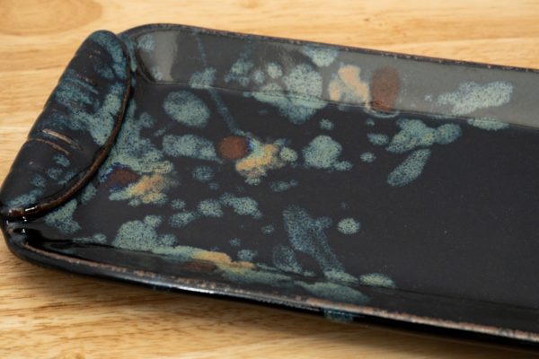 the photograph shows a close up of a clay coyote small tray glazed in midnight garden. the photograph is taken from a close enough point that not all of the small tray is visible. the colorful part of the midnight garden glaze pattern is the focus, and it takes up a majority of the left side. the right side handle is out of the pictures. the small tray is resting on a light colored wooden surface