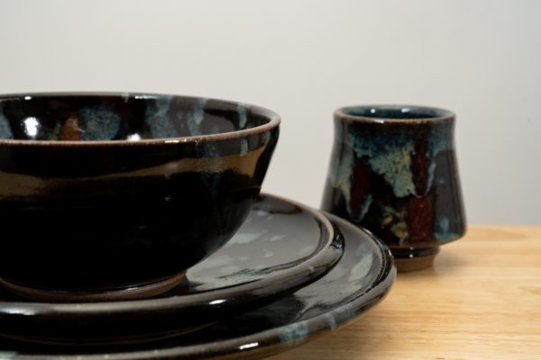 the photograph shows a close up view of a clay coyote tableware place setting. the setting includes 4 pieces, a dinner plate, a sandwich plate a soup and chili bowl and a yunomi cup. the photograph is taken close enough to the pottery that the plates and bowl are not completely in frame. they take up a majority of the lower left of the photograph. the stack goes dinner plate, sandwich plate then bowl on top. the yunomi is slightly behind and to the right of the stack. everything is resting on a small light colored wooden table. the background is a white wall.
