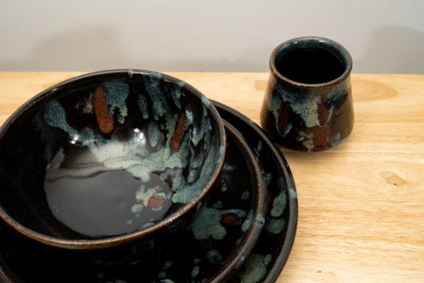 the photograph shows a high angle view of a clay coyote tableware place setting. the setting includes 4 pieces, a dinner plate, a sandwich plate a soup and chili bowl and a yunomi cup. the photograph is taken close enough to the pottery that the plates and bowl are not completely in frame. they take up a majority of the lower left of the photograph. the stack goes dinner plate, sandwich plate then bowl on top. the yunomi is slightly behind and to the right of the stack. everything is resting on a small light colored wooden table. the background is a white wall.