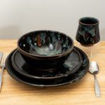 the photograph shows a clay coyote tableware setting. it is 4 pieces of clay coyote pottery, a dinner plate, a sandwich plate, a soup and chili bowl and a yunomi cup. in the photograph the tableware setting pieces are all glazed in midnight garden. there is a fork and a knife on the left hand side of the stack of 2 plates and bowl. on the right side of the stack there is a spoon. in the upper right there is the yunomi on a small clear plastic stand. everything is resting on a small light colored wooden table. the background is a white wall. the photograph is lit with white light.